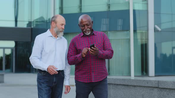 Two Diverse Elderly Colleagues Chatting Outdoors Looking at Screen of Digital Device Using App in