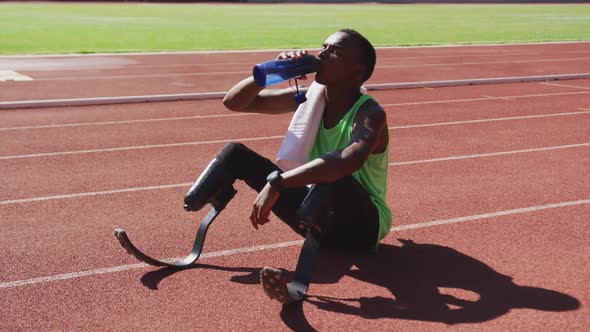 Disabled mixed race man with prosthetic legs sitting on a race track and drinking water
