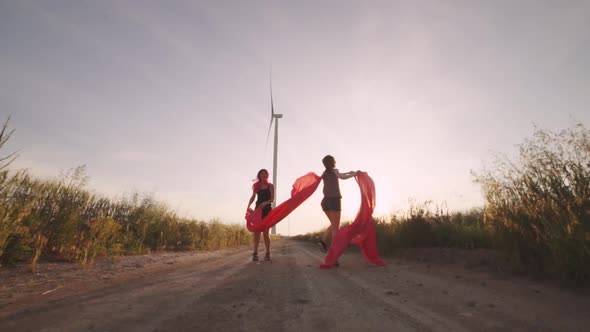 Two Girls with Pieces of Red Cloth Run to the Wind Generator in the Field