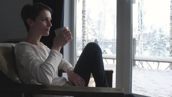 pretty girl sits on chair near window and drinks coffee from Cup.