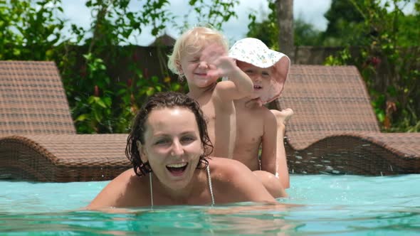 Cheerful Family of Young Mother and Toddler Twin Sisters Swimming in Pool Waving Hello to the Camera