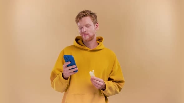 Young Man Eating an Apple and Using a Phone on a Beige Background