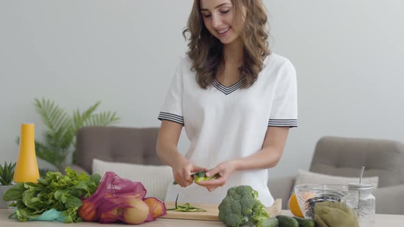 Cute Girl Cuts a Cucumber with Knife While Standing at a Table in the Kitchen at Home