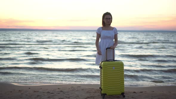 Young Woman with a Yellow Suitcase on the Beach By the Sea. Girl in a White Dress By the Sea at
