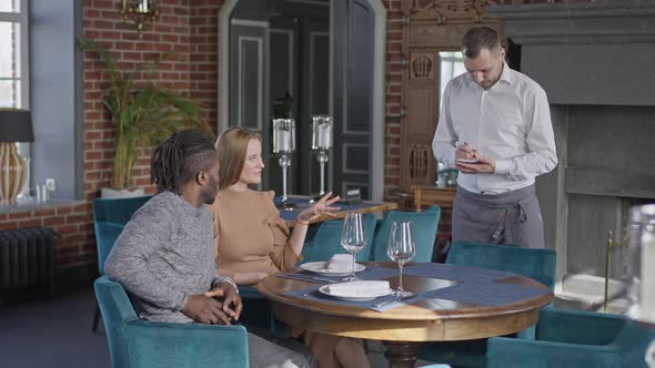 Wide Shot of Male Waiter Taking Order of Wealthy Rich Interracial Couple Dining in Luxurious