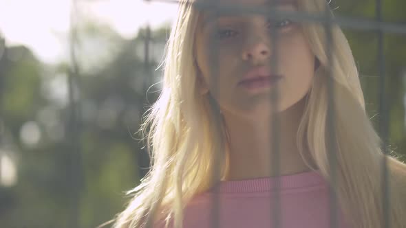 Close-up Face of Charming Blond Woman Standing in Sunrays Behind Mesh Fence. Portrait of Beautiful