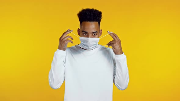 Young African Man Puts on Face Medical Mask During Coronavirus Pandemic. Portrait on Yellow