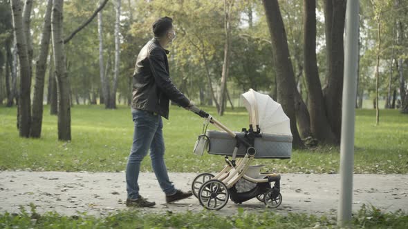 Side View of Young Caucasian Father in Covid Face Mask Pushing Baby Stroller Along Park Alley