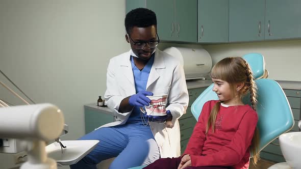 African Dentist Holding Plastic Model of Teeth and Talking to Little Female Patient.