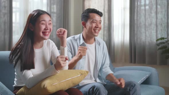 Excited Asian Couple Watching Tv Together Sitting On A Sofa In The Living Room At Home
