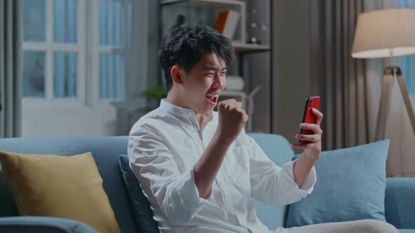 Happy Asian Man Celebrating While Using Smartphone In The Living Room