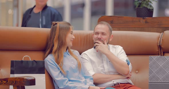 Man and a Girl are Sitting on a Sofa in a Mall and Discussing Something