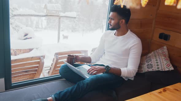 Young Indian Man Relaxing Near the Window in Cozy Home Drinking Coffee and Using Laptop