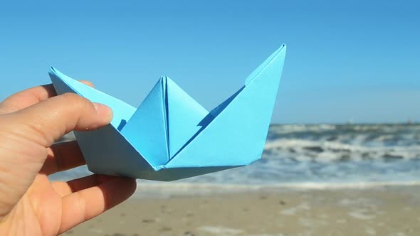 Person Holding Blue Paper Boat in Hand on Background of Sea Waves on Sunny Day