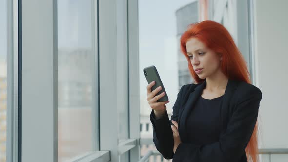 Portrait of a Red-haired Woman Holding a Mobile Phone in Her Hand Looking Gloomily Out the Window