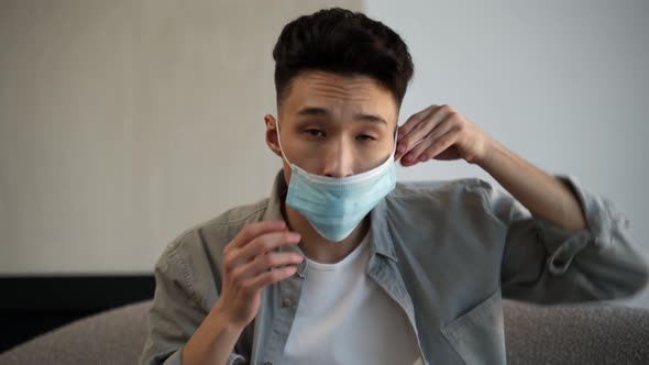 Young Asian Man Puts on Medical Mask on Face Sitting on Sofa