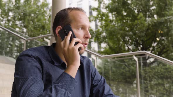 A Caucasian Man Talks on a Smartphone As He Sits on a Staircase in an Urban Area