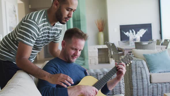 Multi ethnic gay male couple sitting on couch playing guitar together