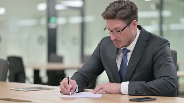 Focused Middle Aged Businessman Writing on Paper in Office