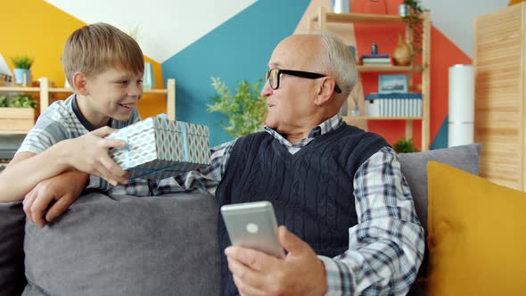 Grandfather Using Smartphone When Grandson Giving Gift Box Congratulating at Home alt
