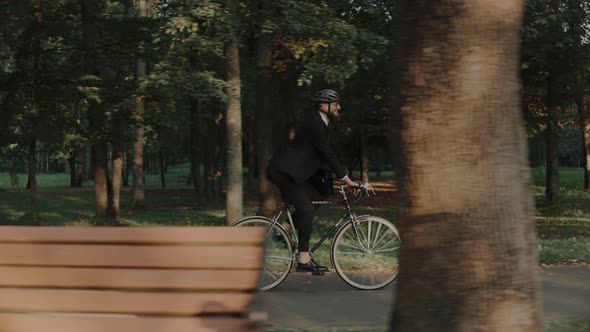 Young Man Riding a Bike in City Park Wearing Business Suit and Helmet Side View