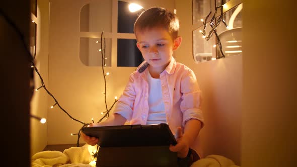 Portrait of Little Boy Watching Cartoons on Tablet Computer at Night While Sitting in Small Toy