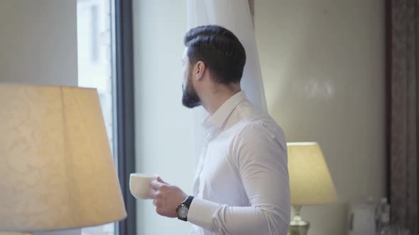 Confident Handsome Businessman Drinking Morning Coffee Next To Window in Hotel Room