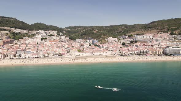 People enjoying sand beach, Sesimbra picturesque townhouses against hills and blue sky.