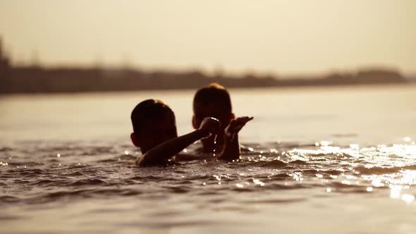 Silhouette of two boys swimming and playing with water at sunset. alt