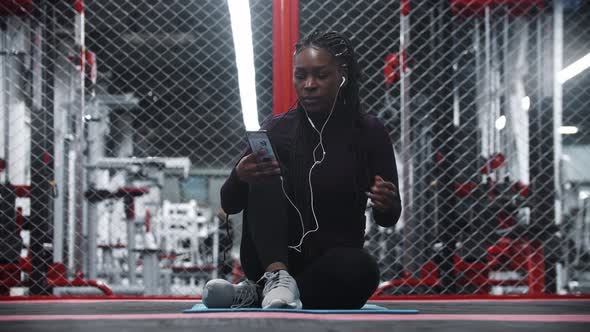 Smiling Africanamerican Woman Sitting on Yoga Mat in a Gym and Taking a Selfie on Her Phone