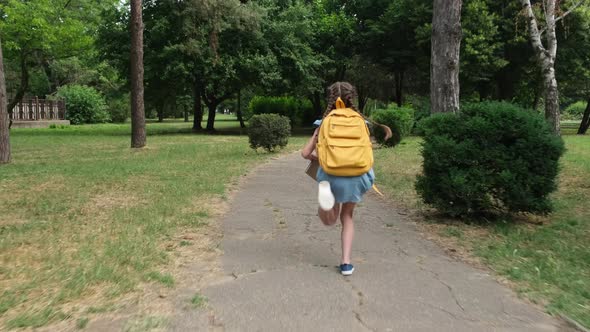 Little Schoolgirl with School Backpack and Book Runs Through the Park to School