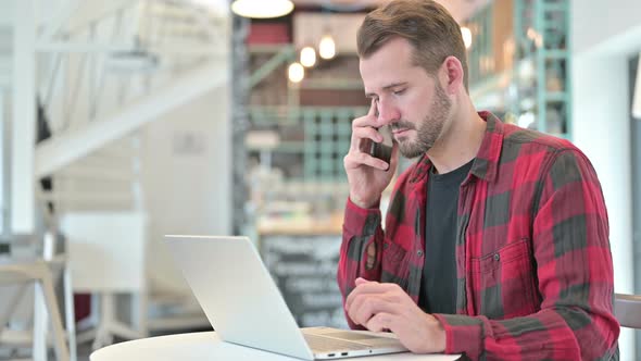 Young Man with Laptop Talking on Smartphone in Cafe