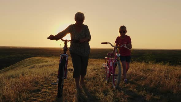 An Elderly Lady Walks with Her Granddaughter Bicycles at Sunset