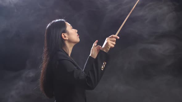 Side View Of Asian Conductor Woman Holding A Baton And Showing Gesture In The Black Studio With Fog