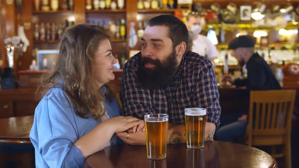 Woman and Man Celebrating Drinking Golden Craft Beer Sitting at Wooden Table Indoors at Pub