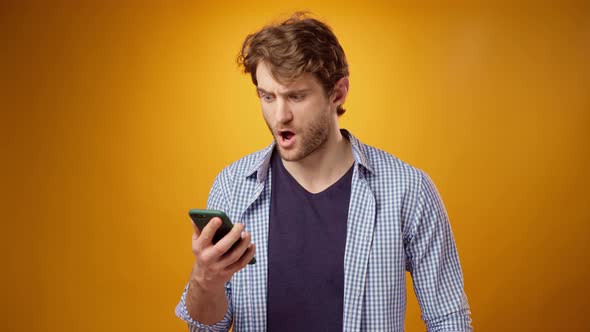 Young Man Looks at Cell Phone Screen with Astonishment and Disappointment Yellow Background