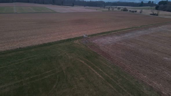 A hunter's tree stand in the middle of green and brown meadows in Germany during sunset. Fast aerial