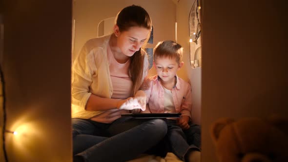 Dolly Shot of Smiling Mother Sitting in Toy House or Tent with Her Little Son and Using Tablet