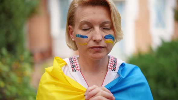 Portrait of Depressed Ukrainian Adult Woman Praying in Slow Motion Outdoors Wrapped in National Flag