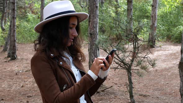 A stylishly dressed woman mixe-race is holding a phone in her hands while walking through the woods.