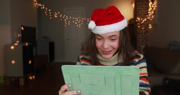 Smiling Brunette in Christmas Hat Looks at Tablet Screen