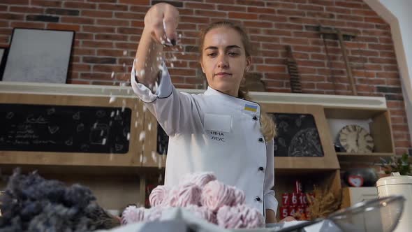 Female Confectioner Sprinkling Powdered Sugar on Pink Marshmallows on the Plate in the Kitchen