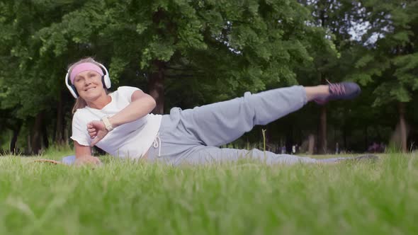 Happy Mature Woman Practicing Yoga and Listening To Music with Headphones in Park.