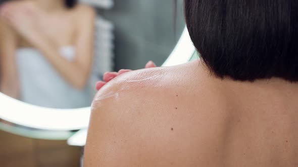 Unknown Brunette Standing in Front of Mirror and Applying Body Cream on Her Shoulder