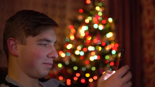 A Young Man Sits with a Tablet in Him Hands on the Background of a Christmas Tree. The Room Is