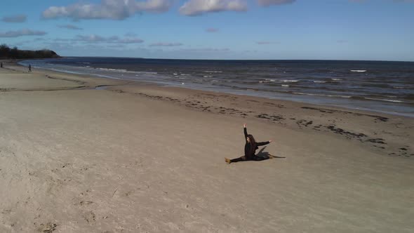 Aerial Fly Around Young Gymnast Woman (Wears Black Sweater and Leggings) Practices Split on Sandy