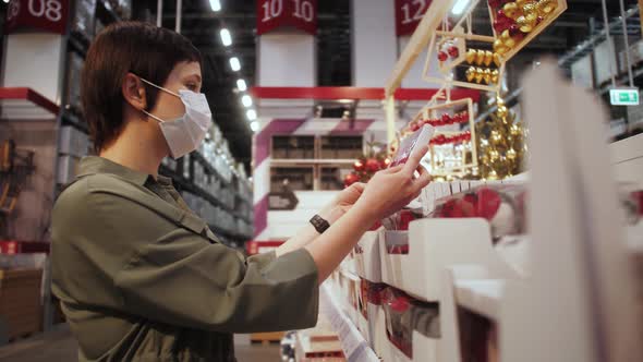 Woman in medical mask looks and buys the Christmas decoration, toys and decorations in mall.