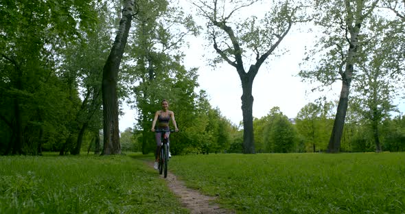 Beautiful Slender Girl in a Tank Top and Leggings Riding a Bike Among the Trees and Green Grass