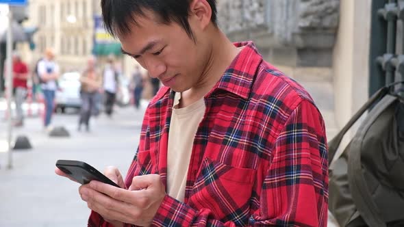 Asian Man Holding Smartphone in His Hands Scrolling Screen with News or Social Media Outdoor