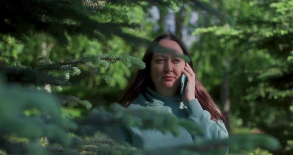 Woman Is Listening Audio Messages In Messenger Standing Behind Fir Tree In Park